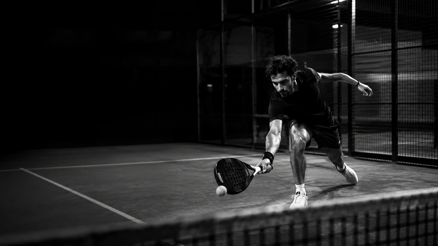 A black and white action photograph of a tennis player lunging to hit a ball with a racket on an indoor court.