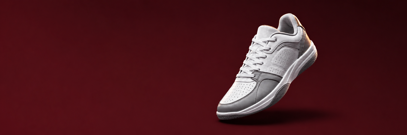 A white athletic sneaker with light gray panels and a subtle gold accent on the heel, shown floating against a solid dark red background.