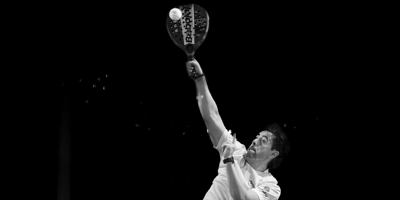 Black and white action photograph of a tennis player mid-serve, holding a tennis racket with a tennis ball above their head against a dark background.