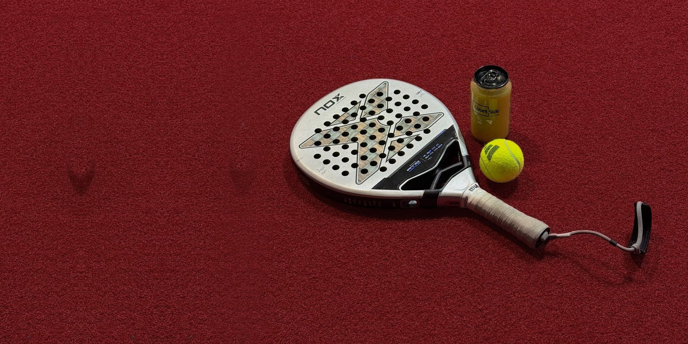 A padel racket with a white and black head, a yellow tennis ball, and a small yellow bottle, arranged on a textured red surface.